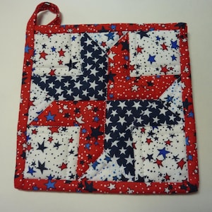May include: Square quilted potholder with a patriotic theme. The design features a patchwork pattern of red, white, and blue fabrics. The white fabric is dotted with red and blue stars. The potholder has a red border and a loop for hanging.