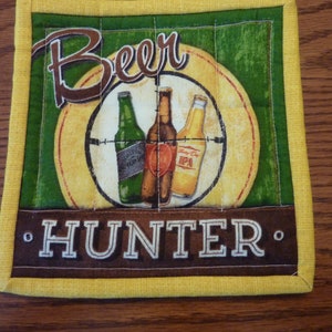 May include: A yellow and green fabric coaster with the words "Beer Hunter" and a graphic of three beer bottles in a target.