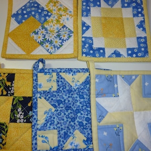 May include: A collection of handmade quilted pot holders in various designs. The pot holders feature a mix of blue, yellow, and white fabrics with floral and geometric patterns. Each pot holder has a loop for hanging.