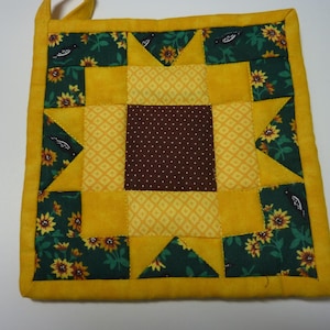 May include: Square quilted pot holder with a sunflower design. The center is brown with white dots, surrounded by yellow squares and triangles. Green fabric with sunflowers and birds accents the design. A yellow loop is attached.