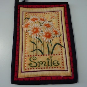 May include: A quilted wall hanging featuring a floral design with white and orange daisies, green stems, and the word "Smile". The border is black with a red and cream accent.