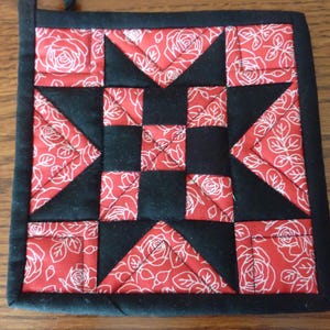 May include: Black and red quilted hot pad with a star design. The fabric has a white floral pattern.
