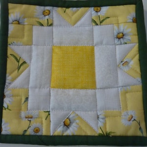 May include: Square quilted pot holder with a daisy pattern. The center is yellow, surrounded by white and yellow squares with daisy accents. A dark green border frames the design.