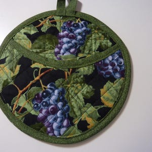 May include: Round quilted pot holder with a green border and hanging loop. The fabric features a pattern of purple grapes and green leaves on a black background. The pot holder is designed for kitchen use.