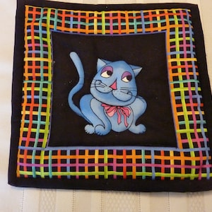 May include: A blue cat with a pink bow sits in the center of a black square with a colorful checkered border. The cat is outlined in black and has white eyes and a white nose.