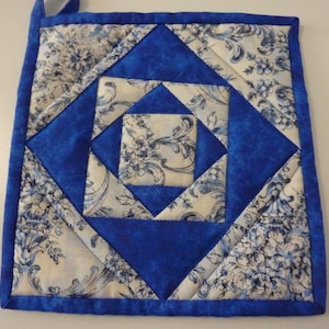 May include: Square quilted pot holder with a blue and white floral pattern. The design features a central square surrounded by concentric diamond shapes in varying shades of blue and white. A blue loop is attached for hanging.