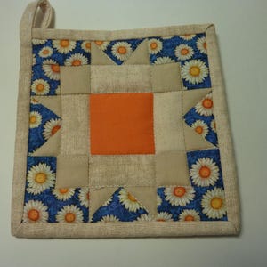 May include: Square quilted pot holder with a loop for hanging. The design features a central orange square, surrounded by tan and blue fabric. The blue fabric is patterned with white daisies.
