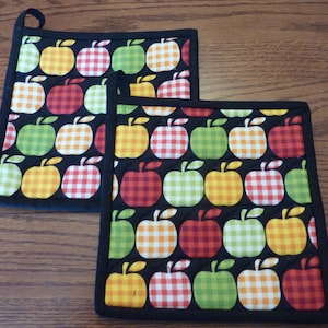 May include: Two black quilted pot holders with a pattern of red, yellow, and green apples on a black background. The apples have a gingham pattern.