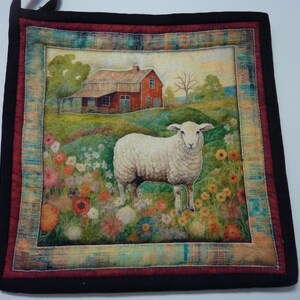 May include: A square quilted potholder featuring a printed scene of a white sheep standing in a field of colorful flowers with a red barn in the background. The design is framed by a decorative border and finished with a solid black fabric binding.