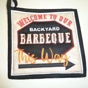 May include: Square quilted pot holder with a black border and a loop for hanging. The center features a sign that reads "WELCOME TO OUR BACKYARD BARBEQUE" with an arrow pointing the way. The text is in red, black, and orange.