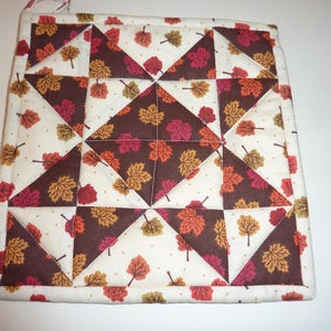 May include: Square quilted potholder with a fall leaf pattern. The design features brown, white, orange, and red triangular and square shapes. The potholder has a loop for hanging and is likely used for handling hot cookware.