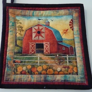 May include: A square quilted potholder featuring a printed scene of a red barn with a star emblem, a white fence, and orange flowers in the foreground. The design is framed by a multi-colored border and finished with a solid black fabric binding.
