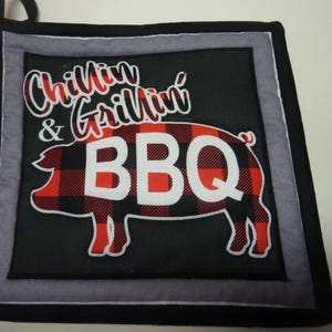 May include: Black square pot holder with a pig graphic in red and black plaid. The words "Chillin' & Grillin' BBQ" are written across the top and inside the pig. The pot holder has a black hanging loop.