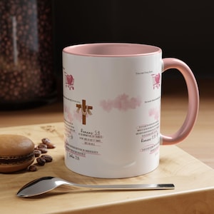 May include: White ceramic mug with a pink interior and handle. The mug features a cross and text from Romans 5:8, along with other scriptural references and pink cloud graphics. A spoon and a macaron are in the foreground.