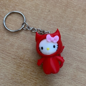 Puede incluir: Llavero Hello Kitty rojo con un disfraz de diablo, un lazo rosa y un llavero plateado.