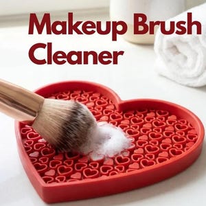 Op de afbeelding: Een rode hartvormige siliconen make-up kwastenreiniger mat met een textuur van kleine verhoogde hartjes. Een make-up kwast met een zwarte steel en roségouden huls reinigt op de mat met wit zeepsop. Tekst leest: Makeup Brush Cleaner.