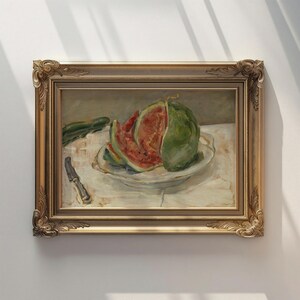 May include: A framed still life painting of a watermelon on a white plate. The watermelon is cut into slices, revealing its red flesh. A knife and a cucumber are also visible. The frame is gold-colored.