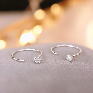 May include: Two open-ended silver rings, each featuring a small, sparkling, round gemstone at one end. The rings are displayed on a soft, gray surface, with a blurred, warm-toned background.