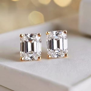 May include: A pair of dazzling stud earrings featuring emerald-cut, clear gemstones set in gold. The earrings are displayed on a white surface, with a soft, blurred background of warm, golden tones.