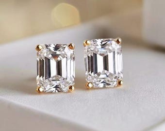 May include: A pair of dazzling stud earrings featuring emerald-cut, clear gemstones set in gold. The earrings are displayed on a white surface, with a soft, blurred background of warm, golden tones.