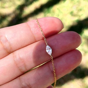 Pu&ograve; includere: Delicato braccialetto con catena dorata con diamante taglio marquise. Il diamante &egrave; il punto focale, incastonato in un design minimalista. Il braccialetto &egrave; presentato su uno sfondo verde sfocato, evidenziando l'eleganza del gioiello.