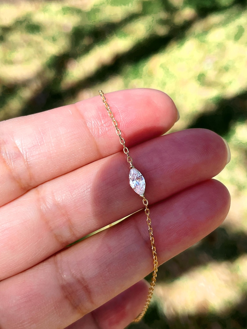 Pu&ograve; includere: Delicato braccialetto con catena dorata con diamante taglio marquise. Il diamante &egrave; il punto focale, incastonato in un design minimalista. Il braccialetto &egrave; presentato su uno sfondo verde sfocato, evidenziando l'eleganza del gioiello.