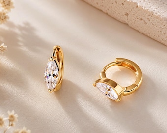 May include: A pair of gold hoop earrings featuring a marquise-cut clear gemstone set in the center of each hoop. The earrings have a polished gold finish and a hinged closure mechanism, showcasing a bright, reflective surface on the stones.