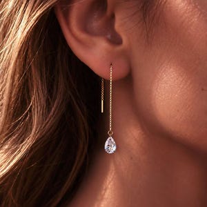 May include: A gold threader earring featuring a delicate box chain that passes through the earlobe, ending in a small, clear, pear-shaped faceted gemstone drop. The earring has a thin, straight gold post at the top of the chain.