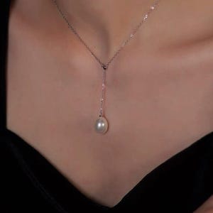 May include: A delicate silver necklace with a single, luminous pearl pendant. The necklace features a fine chain that drapes down to a teardrop-shaped pearl. The pearl is a creamy white color, and the necklace is set against a dark background.