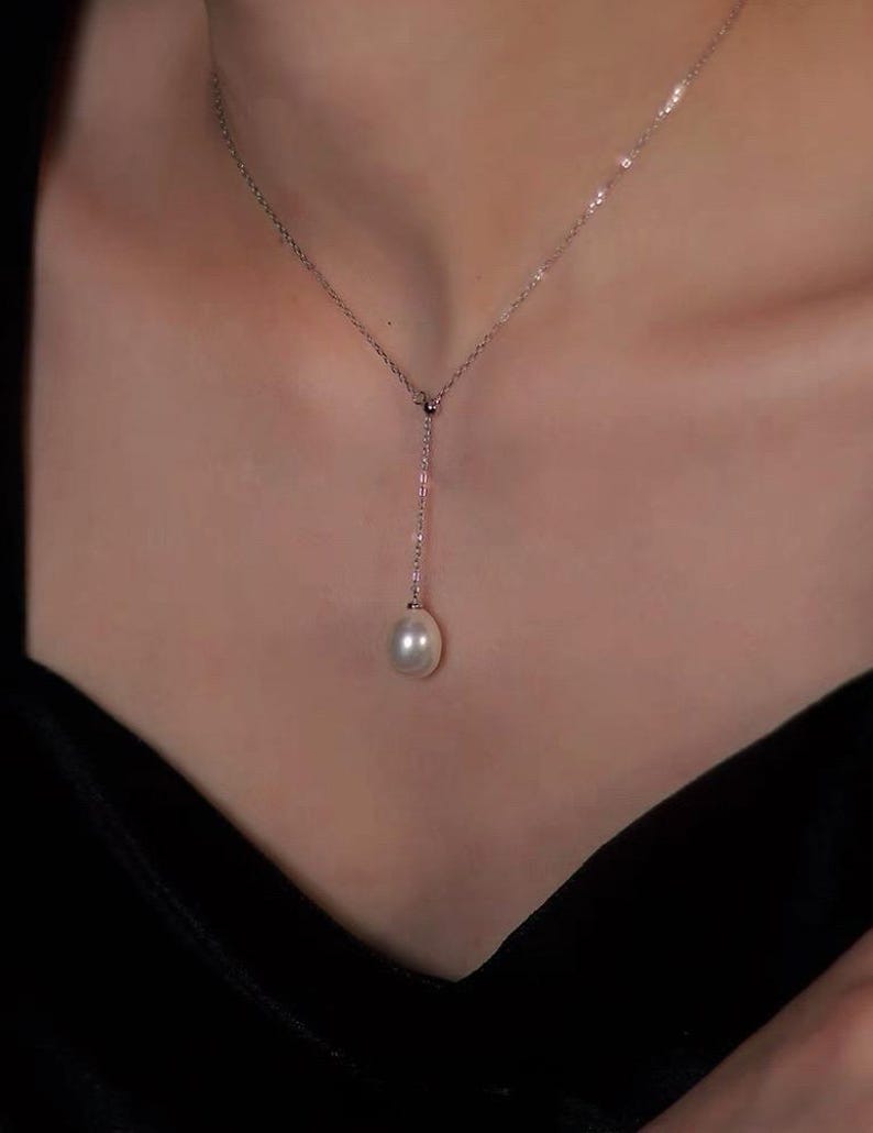 May include: A delicate silver necklace with a single, luminous pearl pendant. The necklace features a fine chain that drapes down to a teardrop-shaped pearl. The pearl is a creamy white color, and the necklace is set against a dark background.