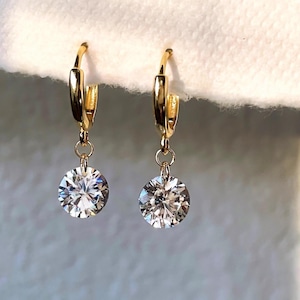 May include: A pair of gold hoop earrings featuring a polished metal finish and a single round, clear, faceted gemstone drop suspended from each hoop. The earrings have a hinged closure mechanism and exhibit a bright, reflective metallic luster.