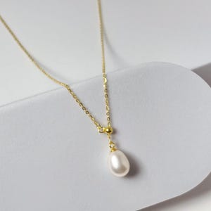 May include: A delicate gold-tone necklace featuring a single, teardrop-shaped pearl pendant. The chain is fine and elegant, with a small bead accent above the pearl. The necklace is displayed on a white, curved surface, highlighting its simple beauty.