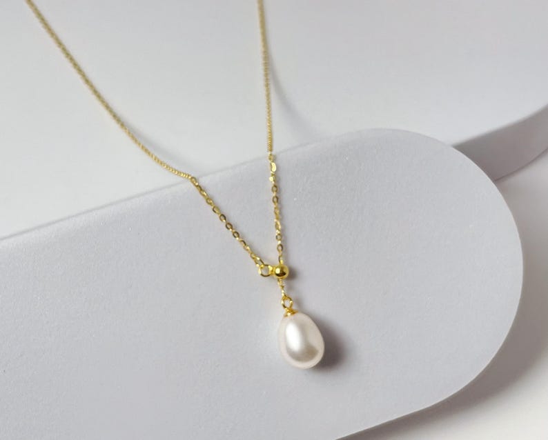 May include: A delicate gold-tone necklace featuring a single, teardrop-shaped pearl pendant. The chain is fine and elegant, with a small bead accent above the pearl. The necklace is displayed on a white, curved surface, highlighting its simple beauty.