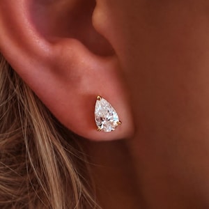 May include: A pear-shaped clear gemstone stud earring set in a gold-toned metal prong setting. The faceted stone is positioned on an earlobe, showcasing a brilliant cut that reflects light with a sharp, pointed top and a rounded base.
