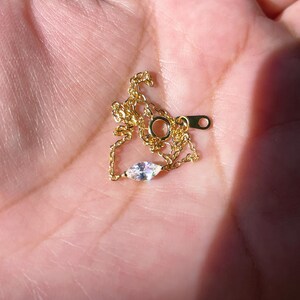 Pu&ograve; includere: Una delicata collana color oro con un piccolo pendente a forma di marquise iridescente. La catena sottile e la chiusura sono visibili. La collana &egrave; esposta su uno sfondo chiaro, probabilmente una mano.