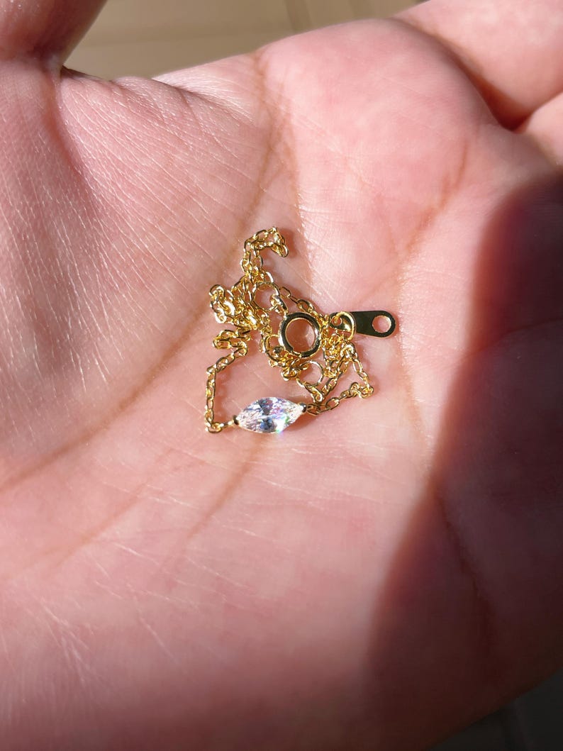 Pu&ograve; includere: Una delicata collana color oro con un piccolo pendente a forma di marquise iridescente. La catena sottile e la chiusura sono visibili. La collana &egrave; esposta su uno sfondo chiaro, probabilmente una mano.