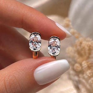 May include: A pair of gold hoop earrings featuring oval-shaped, clear faceted stones set in a bezel setting. The earrings have a polished gold finish and a secure hinged closure, showcasing a classic design with brilliant, reflective center stones.