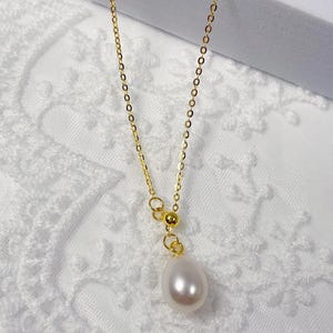 May include: A delicate gold-colored chain necklace with a teardrop-shaped white pearl pendant. The necklace is displayed on a white lace fabric, highlighting the jewelry's elegant design. The chain has a simple, classic style.