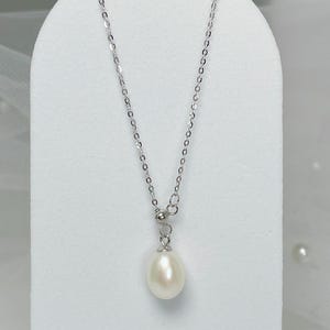 May include: A delicate silver necklace featuring a teardrop-shaped white pearl pendant. The chain is fine and the pearl has a smooth, lustrous surface. The necklace is displayed on a white stand, highlighting its elegant design.