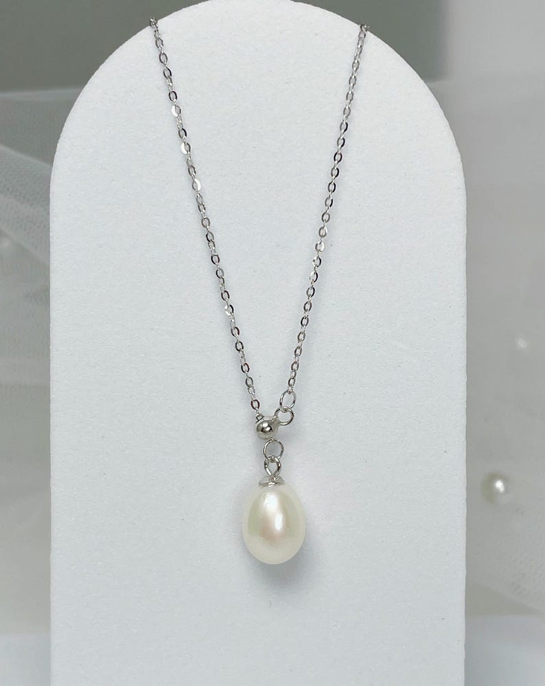May include: A delicate silver necklace featuring a teardrop-shaped white pearl pendant. The chain is fine and the pearl has a smooth, lustrous surface. The necklace is displayed on a white stand, highlighting its elegant design.