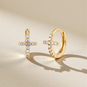 May include: A pair of gold-colored hoop earrings, each featuring a cross design. The crosses and the hoops are embellished with small, clear gemstones. The earrings are displayed on a light beige surface, with soft shadows.