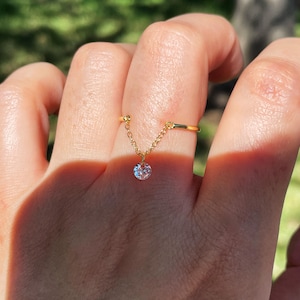 May include: A gold-colored ring with a delicate chain and a round, clear gemstone. The ring is on a finger, with a blurred green background. The chain connects the ring to a small, sparkling pendant.