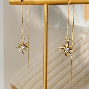 May include: A pair of gold-colored earrings with a starburst design. Each earring features a star-shaped pendant with a central clear gemstone and a delicate chain that dangles below. The earrings are displayed on a gold-colored jewelry stand.