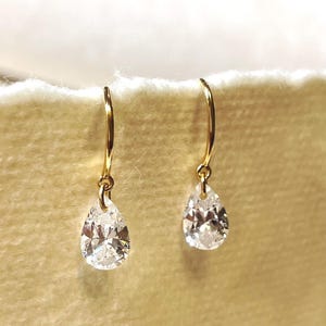 May include: A pair of gold-colored drop earrings with pear-shaped, clear, faceted gemstones. The earrings hang from curved hooks, and the gemstones sparkle with multiple facets. The background is a soft, neutral color.