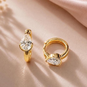 May include: A pair of gold hoop earrings, each featuring a single pear-shaped clear gemstone set at the front. The earrings have a polished metallic finish and a hinged closure mechanism, showcasing a classic design with faceted stones.