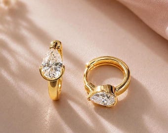 May include: A pair of gold hoop earrings, each featuring a single pear-shaped clear gemstone set at the front. The earrings have a polished metallic finish and a hinged closure mechanism, showcasing a classic design with faceted stones.