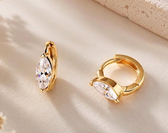 May include: A pair of gold hoop earrings, each featuring a single marquise-cut clear gemstone set at the front. The earrings have a polished gold finish and a hinged closure mechanism, showcasing a bright, reflective surface on the metal and stone.