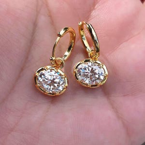 May include: A pair of gold hoop earrings featuring oval-shaped clear faceted stones set in pronged gold settings. Each earring consists of a small circular hoop connected to a dangling oval stone, creating a reflective and polished appearance.