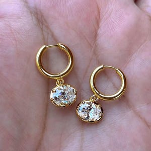 May include: A pair of gold hoop earrings featuring dangling oval-shaped clear gemstones. Each earring consists of a polished gold hoop with a hinged closure and a single faceted oval stone secured in a gold prong setting hanging from the bottom.