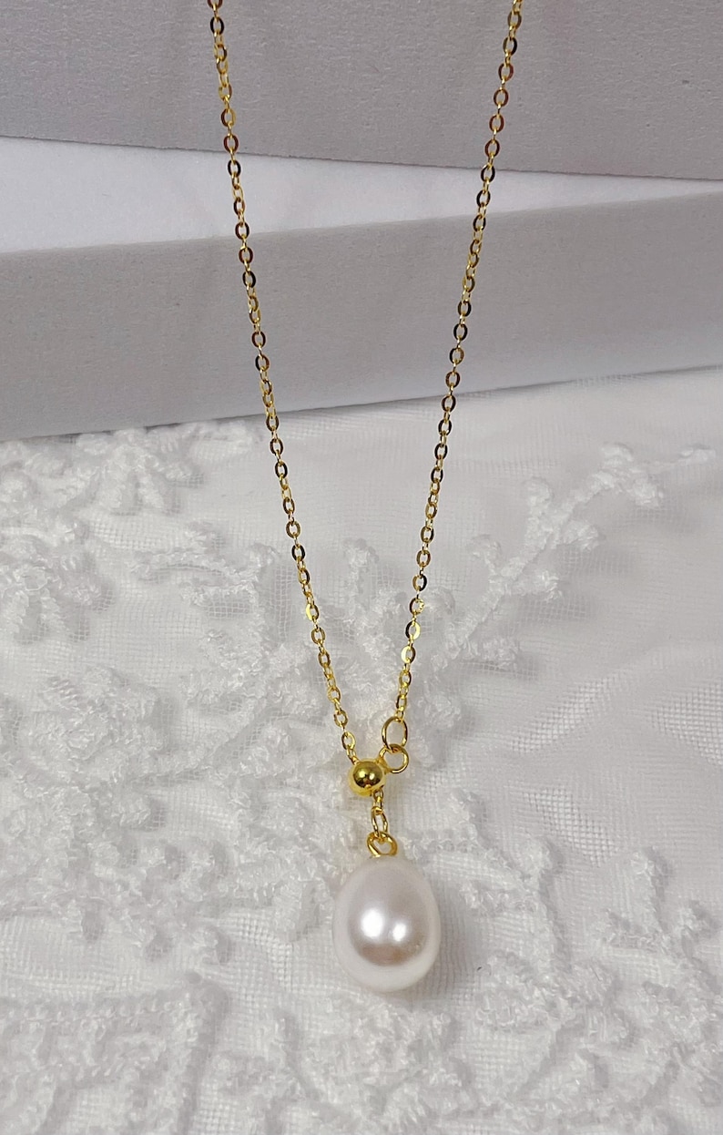 May include: A delicate gold-colored necklace featuring a lustrous, teardrop-shaped pearl pendant. The chain is fine and elegant, with a small gold bead accent. The necklace is displayed against a soft, white background, highlighting its simple beauty.